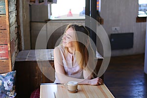 Pensive girl sitting with cup of coffee near table in cafe with nice atmosphere.