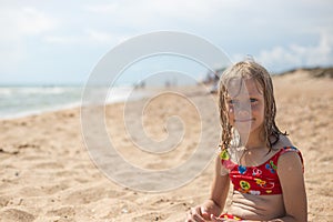 A pensive girl with wet blond hair in a swimsuit sits on the sandy seashore. Summer vacation, travel and tourism