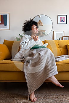 Pensive happy African american woman reading a book at home sitting on sofa relaxing drinking coffee. Vertical image.