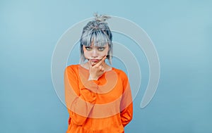 Pensive hipster girl with blue hair and in an orange sweater stands on a blue background and looks at the camera and thinks