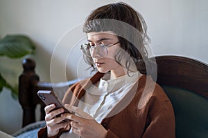 Pensive hipster girl in glasses wearing vintage style looking smartphone screen reading information.