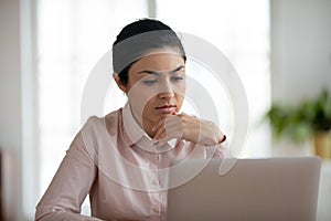 Pensive Indian female employee work on laptop thinking