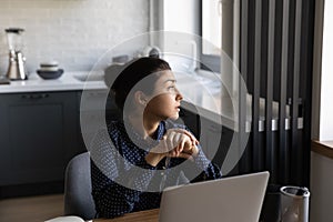Pensive indian female sit by desk indoors look at window
