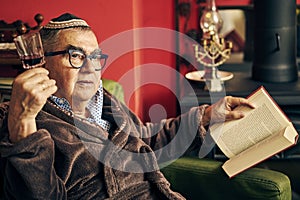 Pensive Jewish senior reading a torah book and drinking kosher wine