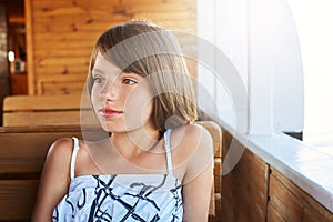 Pensive little child with bobbed hair resting on wooden deck, looking aside while dreaming about something. Pretty girl in white a photo