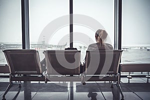 Pensive lonely woman tourist in airport terminal sitting on chair and looking on airplanes through window in departure holding zon