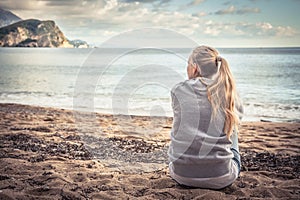Pensive lonely young woman sitting on beach hugging her knees and looking into the distance with hope