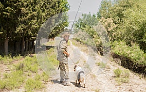 Pensive male fisherman in camouflage outfit walking on dirt road with faithful dog in sunny day.