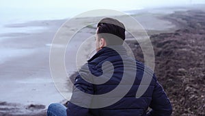 Pensive man sits on bare beach stones at vast grey sea