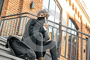 Pensive man sitting on concrete steps and wrapping up in black coat
