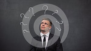 Pensive man in suit looking up, question marks painted on blackboard, solution