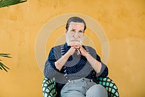 Pensive mature bearded man looking at camera, sitting in chair. Senior male in stylish clothes dreaming about something