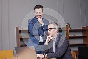 Pensive mature businessman in blue suit and glasses and his smiling colleague are thinking by the laptop in office