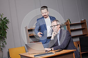 Pensive mature businessman in blue suit and glasses speaking with colleague by the laptop in office