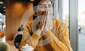 Pensive middle age man in yellow sweater seat by window, hands clasped in contemplation, with reflection in glass. Tired