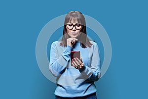 Pensive middle aged woman reading on smartphone, blue background