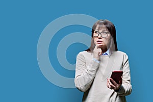 Pensive middle aged woman with smartphone, blue background