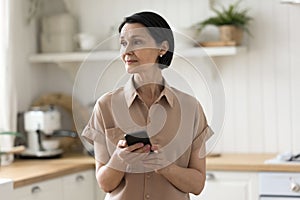 Pensive middle-aged woman standing in kitchen with smartphone