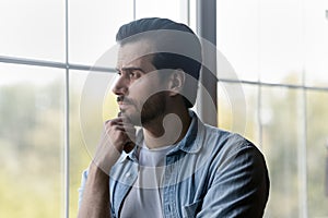 Pensive millennial bearded guy stand by window look at distance