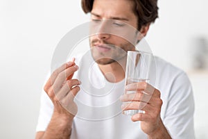 Pensive millennial european man hold glass of water and look at pill in hand