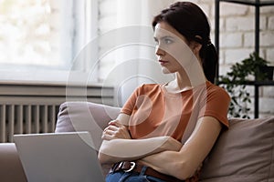 Pensive millennial lady sitting on couch using laptop planning work