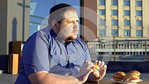 Pensive obese man eating burger, hesitating to start new life, weight loss