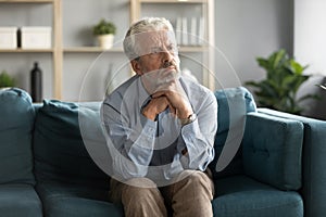 Pensive old 50s man seated on couch feels lonely