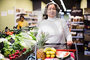Pensive older woman walking with shopping cart at store