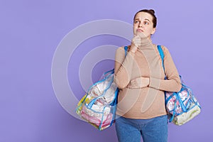 Pensive pregnant woman dreaming about child, posing with bags full of stuff for maternity house, keeps hand under chin, looking