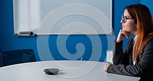 Pensive red-haired office manager in glasses and a suit in an empty conference room. Business woman waiting for