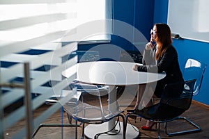 Pensive red-haired office manager in glasses and a suit in an empty conference room. Business woman waiting for