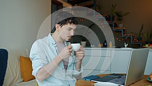 Pensive remote worker drinking coffee and looking at laptop screen at kitchen table at home