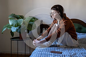 Pensive sad young woman sits on bed, hugs knees next to mobile phone waiting for call, message, sms