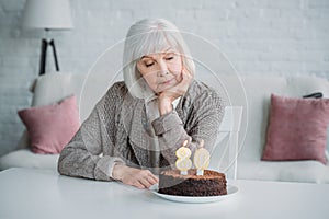 pensive senior lady sitting at table with birthday cake with candles alone