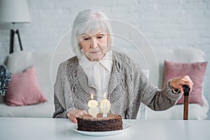 pensive senior lady sitting at table with birthday cake with candles alone