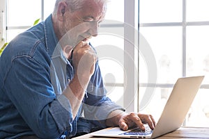 Pensive senior man, with a white beard, smiles looking at his laptop. An elderly Caucasian person. Elderly person and new