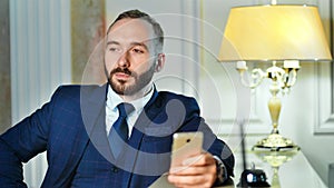 Pensive smiling male businessman writing message using smartphone near reception desk at hotel