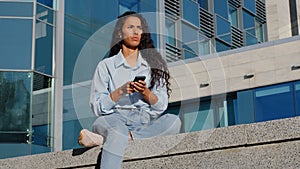 Pensive stressed young woman girl with mobile phone sits in city near building thinks about problem looking for