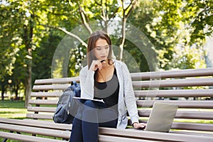 Pensive student girl sitting in park using laptop