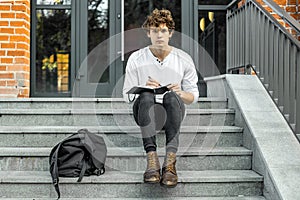 Pensive student sitting on modern building steps and making notes in notepad.
