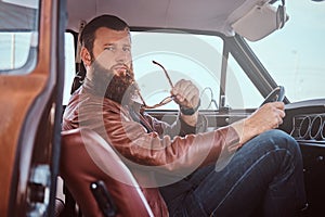 Pensive stylish male holds sunglasses dressed in old-fashioned clothes sits behind the wheel of a tuned retro car with