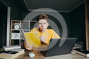 Pensive teenage boy sitting at a table at home with a laptop and books with a notebook and a pen in his hands, looking at camera