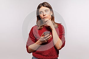 Pensive thoughtful woman in shaggy sweater standing with mobile phone, touching chin and thinking over message