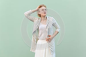 Pensive thoughtful woman wearing striped shirt and skirt, keeps hand on head, looking away, thinking