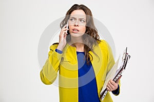 Pensive unpleased young woman with clipboad talking on mobile phone