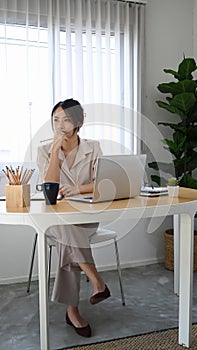 Pensive woman entrepreneur sitting at her workplace and looking away thinking future project.