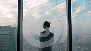 Pensive woman looking at cityscape through window of skyscraper - back view