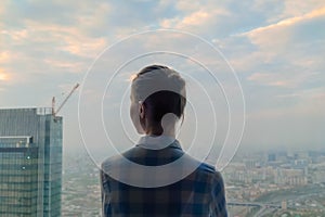 Pensive woman looking at cityscape through window of skyscraper - back view