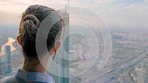 Pensive woman looking at cityscape through window of skyscraper - back view