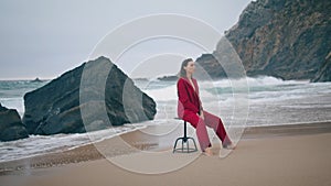 Pensive woman sitting beach in red suit cloudy day. Girl posing chair at shore